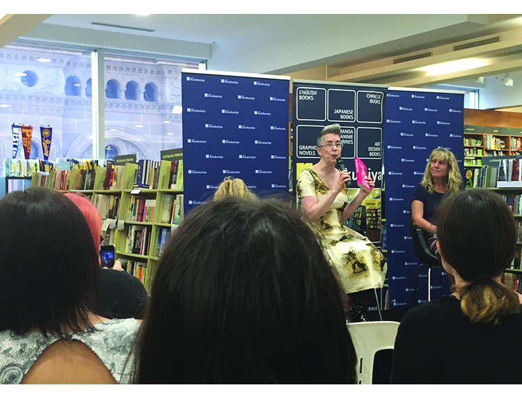 in the background there are many shelves of books, in the forground you can see the back of peoples' heads. In the middle, you can see two white women seated in front of a Kinokunyia banner, one is holding up a copy of the book Summer Skin