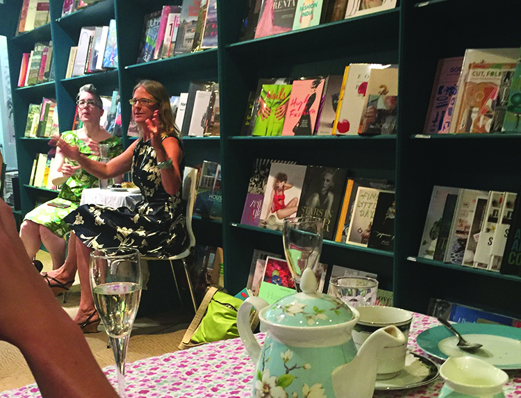 Photo of two white women seated in front of a wall of books gesturing as they speak.