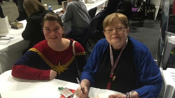 Photo with Tamora Pierce. Shows two white women behind a table, one has brown hair and is wearing a red, and blue Captain Marvel jumper. One has short blondish hair and glasses and is wearing blue. She is holding pen to sign books.