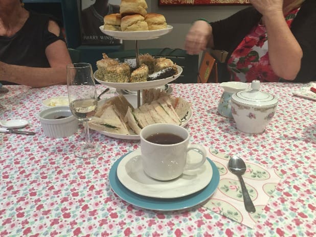 Photograph of a table with a cup of tea in a saucer in front of a three tier cake stand with high tea: cakes, scones and sandwiches